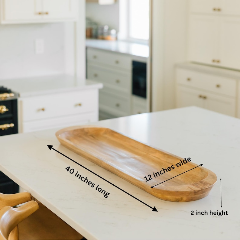 May include: A wooden serving tray, 40 inches long, 12 inches wide, and 2 inches high. The tray is a light brown color and has a long, oval shape. It is sitting on a white countertop in a kitchen setting.