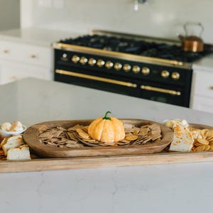 May include: A wooden serving tray with three sections, filled with crackers, cheese, and a small pumpkin. The tray is on a white countertop in front of a black and gold stove.