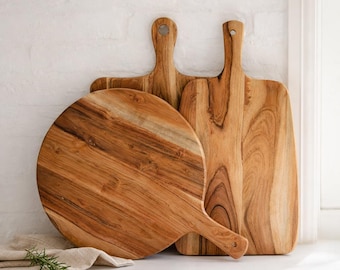 May include: A set of three wooden serving boards featuring natural brown wood grain patterns. The collection includes one circular board and two rectangular boards, each equipped with a handle featuring a hole for hanging storage.
