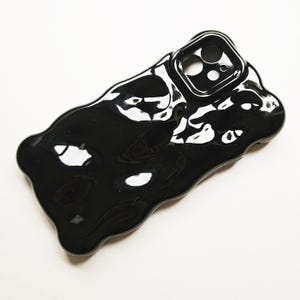 Puede incluir: Carcasa de teléfono negra con un diseño único, abstracto y líquido. La carcasa tiene un acabado brillante y una protuberancia de cámara elevada.