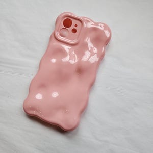 May include: A pink phone case with a textured, melted look. The case has a cutout for the camera lens.