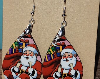 May include: A pair of dangle earrings featuring a teardrop-shaped design with a colorful image of Santa Claus holding a scroll and a bag of presents. The earrings are made of metal with silver hooks.