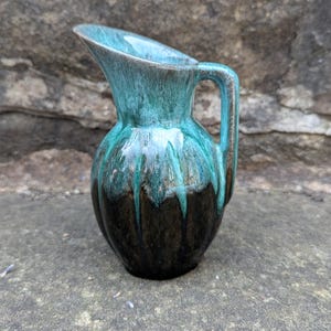 May include: A small, turquoise and brown ceramic pitcher with a handle. The pitcher has a glossy finish with a dripped glaze effect. The pitcher is sitting on a stone surface.