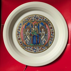 May include: A white decorative plate featuring a detailed, colourful illustration at the centre. The illustration depicts a religious scene with figures, angels, and ornate architectural elements. The plate has a ridged edge.