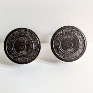 May include: Two black, round, 5 amp fused electrical plugs. Each plug has a raised, circular design with the word "FUSED" and the number "5" in white lettering, surrounded by the word "AMP" in white lettering.