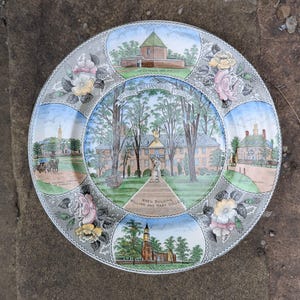 Może przedstawiać: Ozdobny talerz z ilustracjami zabytków historycznego Williamsburg w Wirginii. Talerz ma szarą obwódkę z kwiatowymi wzorami i zawiera obrazy budynków oraz napis "Historic Williamsburg Virginia."