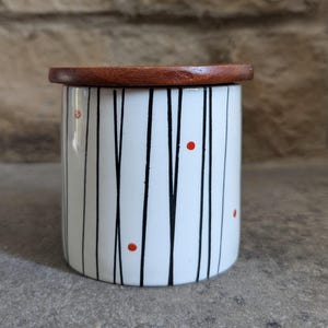 May include: A cylindrical ceramic container with a wooden lid. The white container features vertical black lines and red dots. The lid is a dark brown, circular shape. The container is likely used for storage.
