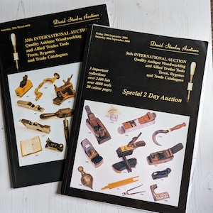 May include: Two black and gold brochures for a 2-day auction of antique woodworking tools, treen, and bygones. The brochures feature images of various tools, including planes, chisels, and a plumb bob. The text on the brochures includes the dates of the auction, the number of lots and tools, and the number of colour pages.