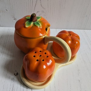 May include: Orange ceramic pumpkin-shaped salt and pepper shakers with a matching creamer. The shakers have a textured surface and the creamer has a lid with a small hole for pouring.
