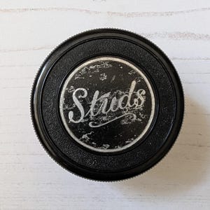 May include: A black, circular object with the word "Studs" in a stylized, cursive font. The text is in a silver color and is set against a distressed, silver background. The outer rim is black and textured.