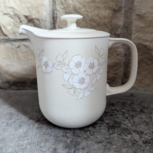 May include: Cream-colored pitcher with a lid and handle. The pitcher features a floral design in light gray. The spout is on the left side, and the handle is on the right. The pitcher is sitting on a gray surface.