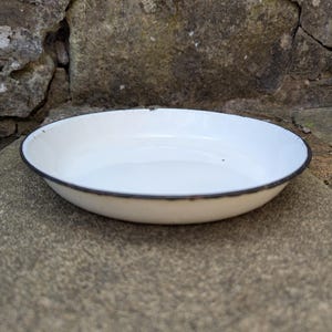 May include: A shallow, round, white enamel bowl with a thin, dark rim. The bowl features a smooth, glossy interior surface and a slightly curved edge, presenting a simple, functional design with visible signs of wear along the outer rim.