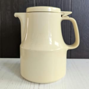 Peut inclure: Une carafe en céramique de couleur crème avec un couvercle et une anse. La carafe a une forme ronde et lisse et une surface légèrement texturée.