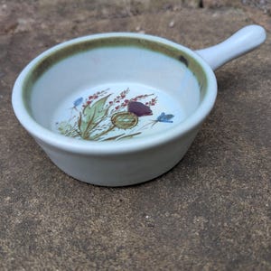 May include: A small, off-white ceramic bowl with a handle. The rim has a brown band, and the interior features a floral design with red, green, and blue accents. The bowl is likely for serving individual portions.