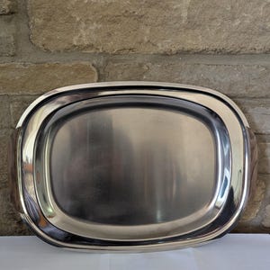 May include: A stainless steel oval serving tray with a polished finish. The tray has a raised rim and integrated handles on each end. The tray is designed for serving food and beverages.