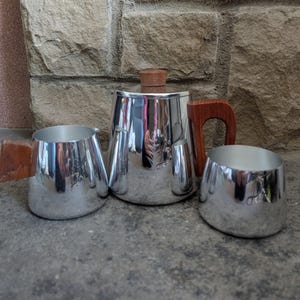 Könnte beinhalten: Ein silbernes Vintage-Teeservice mit Teekanne, Sahnekännchen und Zuckerdose. Die Teekanne hat einen Holzgriff und Deckel. Das Sahnekännchen und die Zuckerdose haben ebenfalls Holzgriffe. Das Set wird auf einer Steinoberfläche präsentiert.