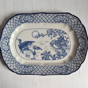 May include: A white oval serving platter with a blue and white floral design. The design features two cranes, a pagoda, and a moon. The border of the platter is a repeating pattern of blue flowers.