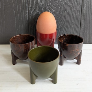 May include: Four egg cups, two brown, one green, and one red, with a single brown egg in the red egg cup.