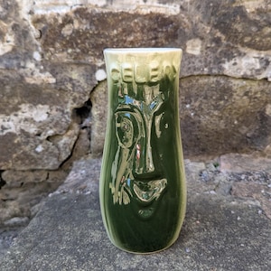 Może przedstawiać: Zielony ceramiczny kubek tiki z rzeźbionym wzorem twarzy. Kubek ma błyszczące wykończenie i jasnozieloną obwódkę. Słowo "CELEBRATE" jest wytłoczone na górze. Kubek jest prezentowany na kamiennej powierzchni.