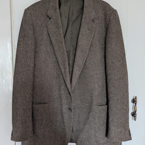 Op de afbeelding: Bruine tweed blazer met een klassiek design, met een gekerfde revers, twee voorzakken en een sluiting met één knoop. De jas hangt aan een hanger tegen een witte deur. De stof van de blazer heeft een subtiel visgraatpatroon.