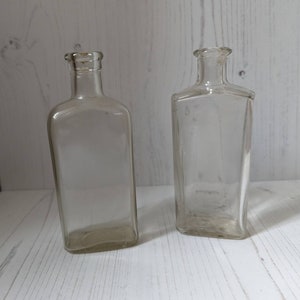 May include: Two clear glass bottles with a rectangular shape. The bottles are empty and have a vintage look.