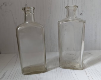 May include: Two clear glass bottles with a rectangular shape. The bottles are empty and have a vintage look.