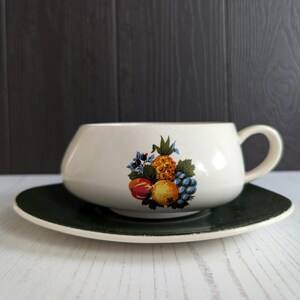 May include: Cream-colored teacup and saucer set. The teacup features a handle and a colorful fruit design including a pineapple, grapes, and peaches. The saucer is dark green with a white rim. A vintage-style tea set.