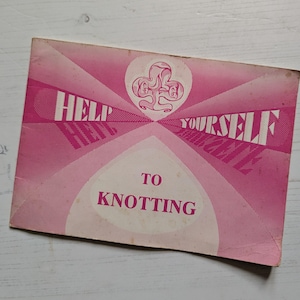 Op de afbeelding: Een vintage roze brochure met de tekst "HELP YOURSELF TO KNOTTING" in het wit. De omslag heeft een geometrisch ontwerp met een centraal embleem. De brochure vertoont tekenen van slijtage.