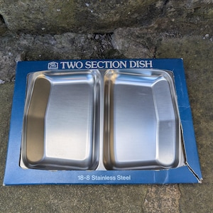 May include: A two-section stainless steel dish in a blue cardboard box. The dish has two rectangular compartments. The box reads "TWO SECTION DISH" and "18-8 Stainless Steel". The dish is on a stone surface.