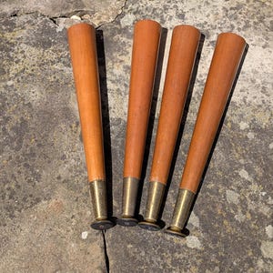 May include: A set of four tapered furniture legs made of light brown wood. Each leg features a smooth, cylindrical wooden body that narrows toward the bottom, connected to a metallic brass-toned ferrule and a small black circular base pad.