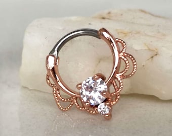 Pode incluir: Joia corporal de ouro rosa com um design delicado e adornado com um cristal transparente. A joia tem a forma de um aro com um pingente.