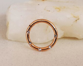 Pode incluir: Um anel de segmento articulado em ouro rosa com seis pedras preciosas claras, redondas e brilhantes. A joia é circular e projetada para piercing corporal. O anel é exibido em uma superfície branca texturizada.