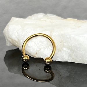 May include: Gold-toned circular barbell with rounded ends. The barbell is on a white surface.