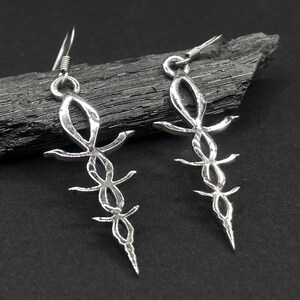 May include: A pair of silver-colored earrings with an ankh design. The earrings have a hook closure and a detailed, openwork design. The ankh symbol is a cross with a loop at the top, a symbol of life. The earrings are displayed on a dark, textured surface.