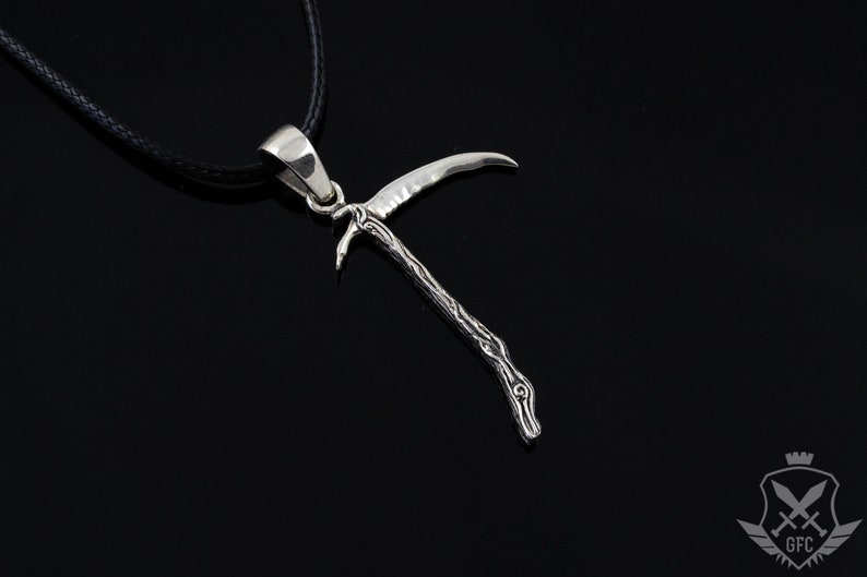 May include: A silver pendant necklace featuring a detailed, stylized depiction of a scythe with a twisted handle. The pendant is hanging from a black cord.