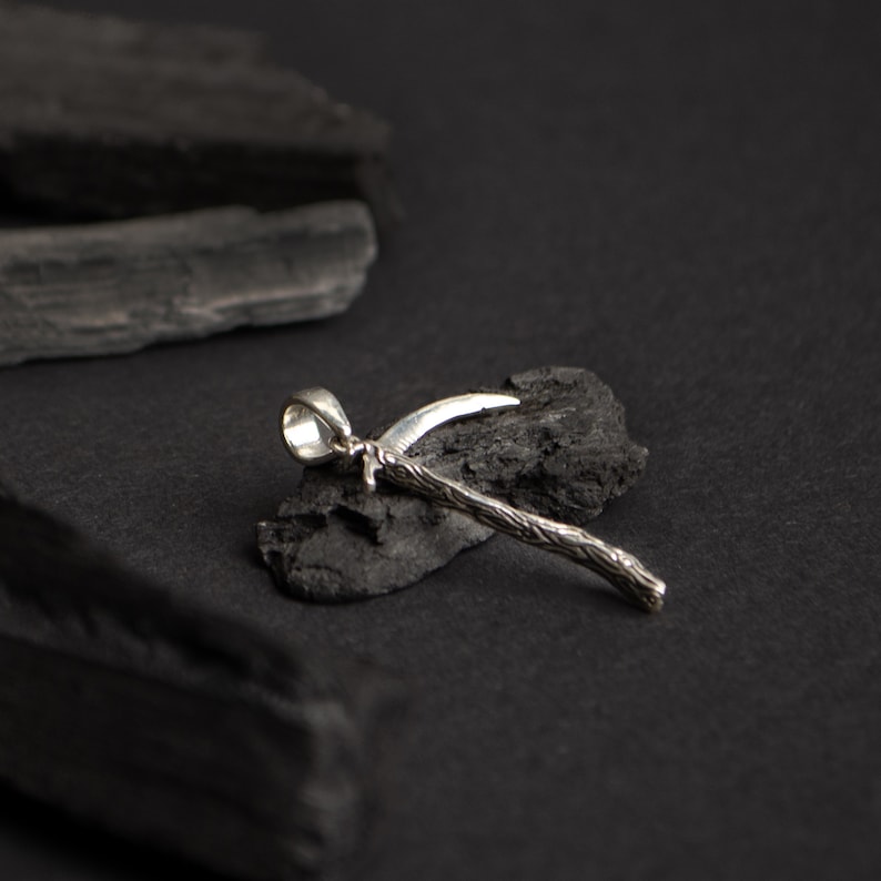 May include: A silver pendant in the shape of a stylized sickle, with a textured handle, resting on a black surface.