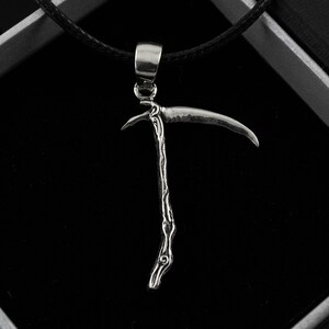 May include: A silver pendant necklace featuring a detailed, stylized depiction of a scythe. The pendant is hanging from a black cord.