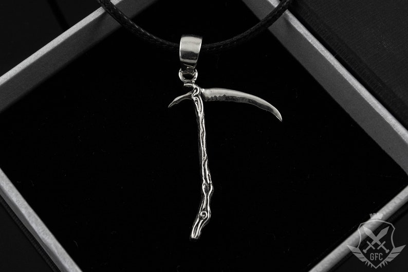 May include: A silver pendant necklace featuring a detailed, stylized depiction of a scythe. The pendant is hanging from a black cord.