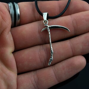 May include: A silver-toned pendant necklace featuring a detailed scythe design. The pendant is hanging from a black cord.