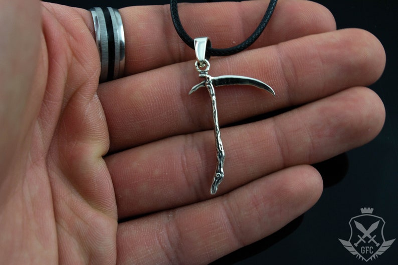 May include: A silver-toned pendant necklace featuring a detailed scythe design. The pendant is hanging from a black cord.