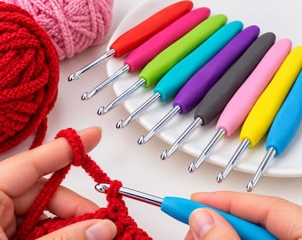 Può includere: Un set di nove uncinetti con manici ergonomici colorati in rosso, rosa, verde lime, turchese, viola, grigio, rosa chiaro, giallo e blu, disposti su un piatto bianco accanto a gomitoli di lana rossa e rosa, con un uncinetto blu in uso.