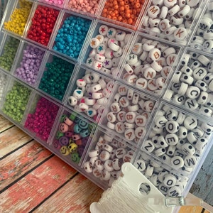 May include: A clear plastic storage container filled with an assortment of small colourful seed beads, white alphabet letter beads, star-shaped beads, heart-shaped beads, and smiley face beads, accompanied by a spool of clear elastic string.