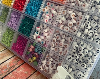 May include: A clear plastic storage container filled with an assortment of small colorful seed beads, white alphabet letter beads, star-shaped beads, heart-shaped beads, and smiley face beads, accompanied by a spool of clear elastic string.