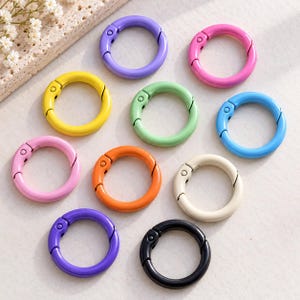 May include: A collection of ten circular spring-loaded metal rings in various colours including purple, pink, yellow, light green, blue, orange, cream, and black. Each ring features a hinged opening mechanism for attaching items securely.