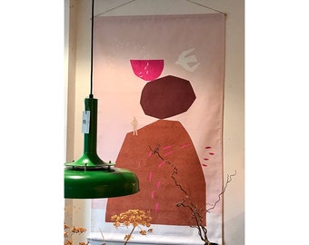 May include: A pink and brown abstract wall tapestry with a white bird and a small figure standing on a rock. The tapestry is hanging on a wall with a green lamp hanging in the foreground.