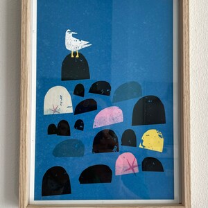 May include: A framed print of a white seagull perched on a black rock with a blue background. The seagull is surrounded by a cluster of colorful rocks in shades of pink, yellow, and black.