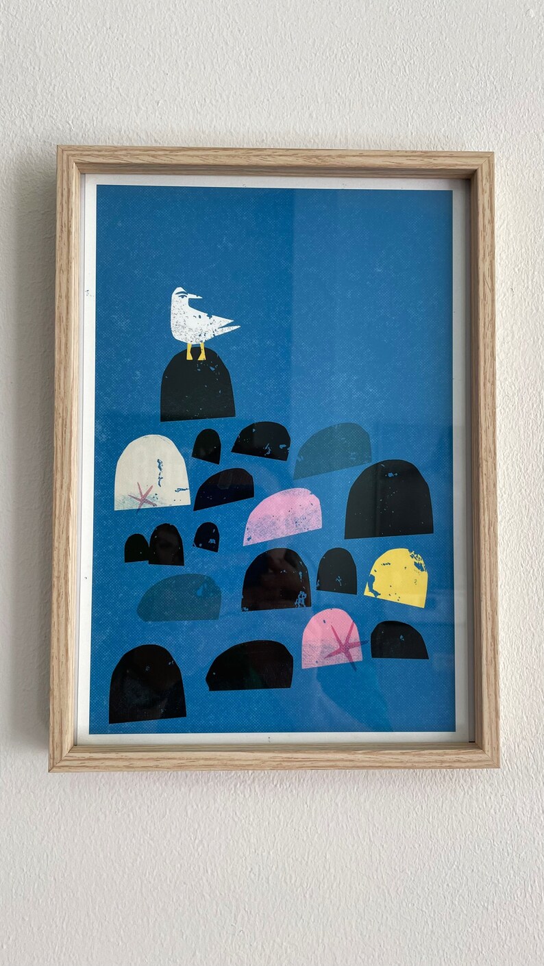 May include: A framed print of a white seagull perched on a black rock with a blue background. The seagull is surrounded by a cluster of colorful rocks in shades of pink, yellow, and black.