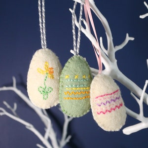 Puede incluir: Tres huevos de Pascua de fieltro colgando de una rama blanca. Los huevos están decorados con bordados coloridos. Un huevo tiene una flor amarilla, otro tiene rayas verdes y amarillas, y el último tiene rayas rosas y moradas.