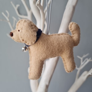 Könnte beinhalten: Ein beigefarbener Filzhund-Anhänger mit einem blauen Filzkragen und einer silbernen Glocke. Der Anhänger hängt an einem weißen Zweig.
