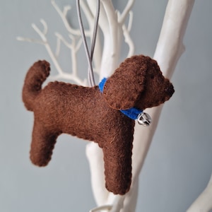 May include: Brown felt dog ornament with a blue collar and a bell hanging from it. The ornament is hanging from a gray string.